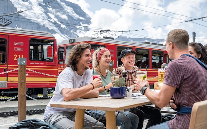 Friends enjoying drinks at Jungfraujoch railway station with red train in background.