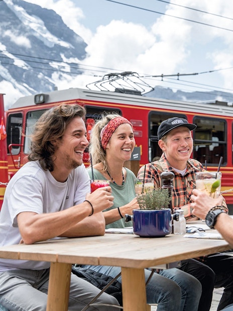 Friends enjoying drinks at Jungfraujoch railway station with red train in background.