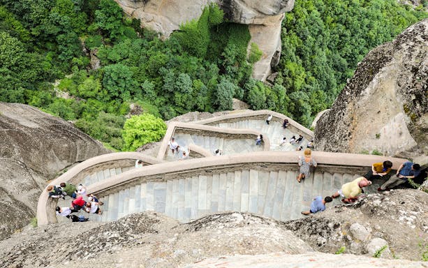 Tourists walking on the stone stairs of the Monastery of Varlaam, surrounded by lush greenery.