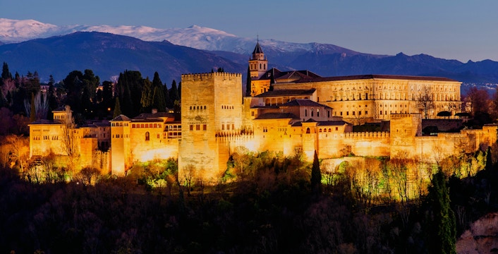 Alhambra palace illuminated at night in Granada, Spain, with mountains in the background.
