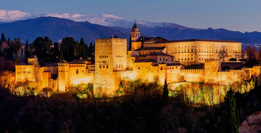 Alhambra palace illuminated at night in Granada, Spain, with mountains in the background.