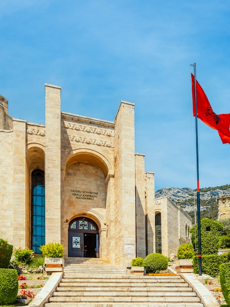 Castle Kruje entrance with Skanderbeg Museum, Kruje, Albania, featuring Albanian flag and mountain backdrop.