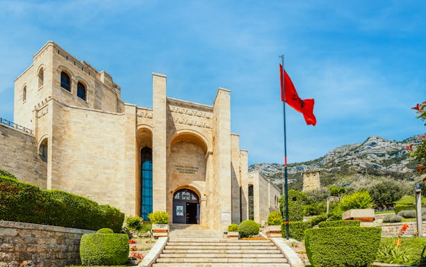 Castle Kruje entrance with Skanderbeg Museum, Kruje, Albania, featuring Albanian flag and mountain backdrop.