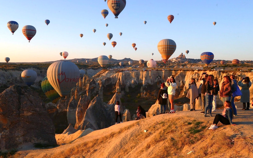 Hot air balloons at sunrise over Cappadocia's fairy chimneys with travelers on a rocky viewpoint.