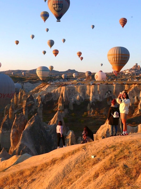 Hot air balloons at sunrise over Cappadocia's fairy chimneys with travelers on a rocky viewpoint.