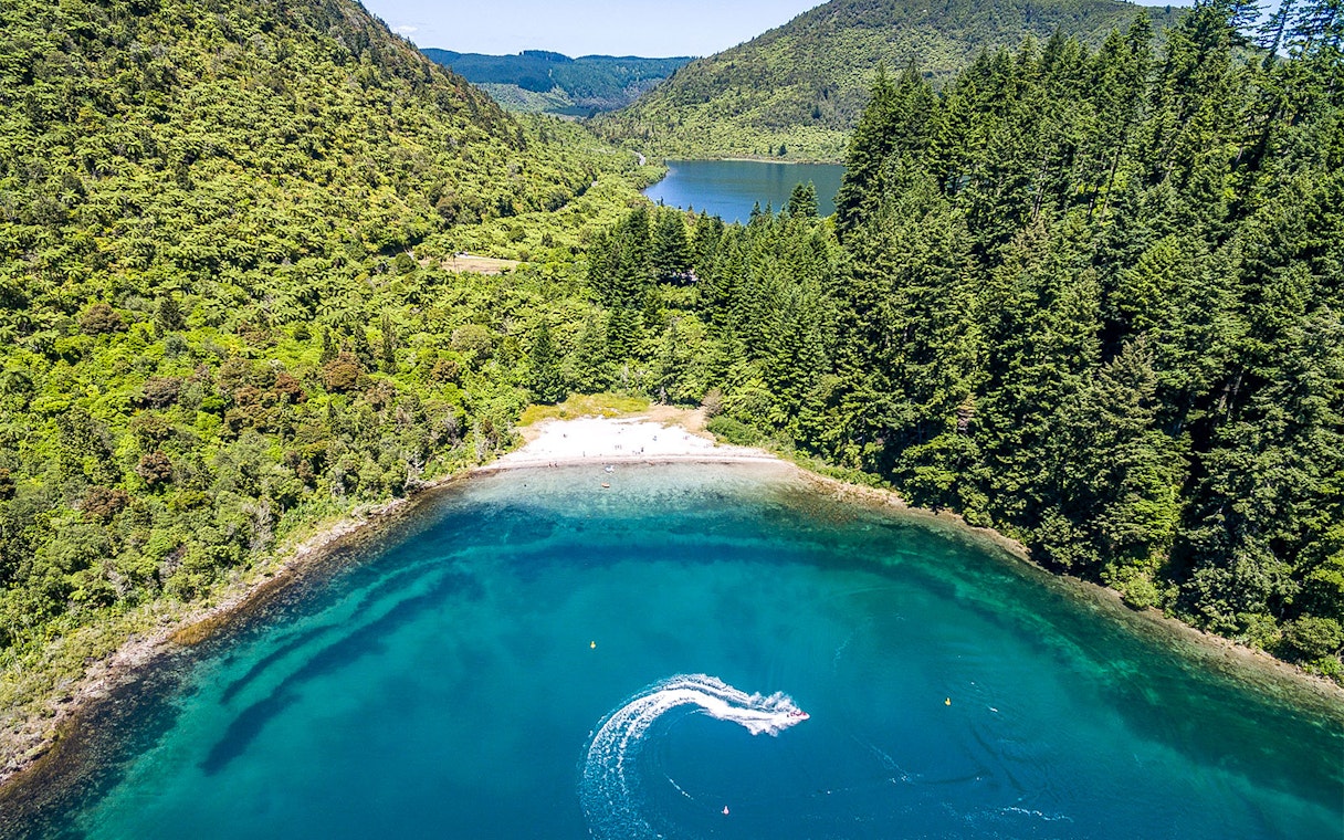 Aerial view of Lake Tarawera with lush green forest and a boat creating a wake in the water.
