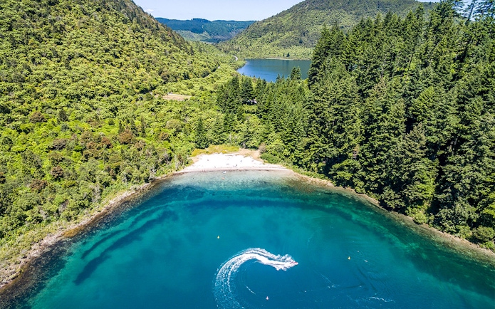 Aerial view of Lake Tarawera with lush green forest and a boat creating a wake in the water.