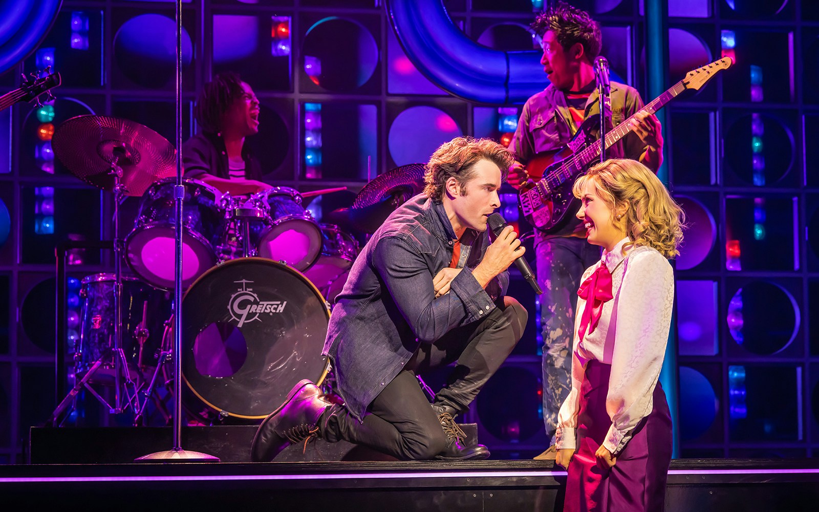 Performer singing to a woman on stage with a band in "The Heart of Rock and Roll" musical.