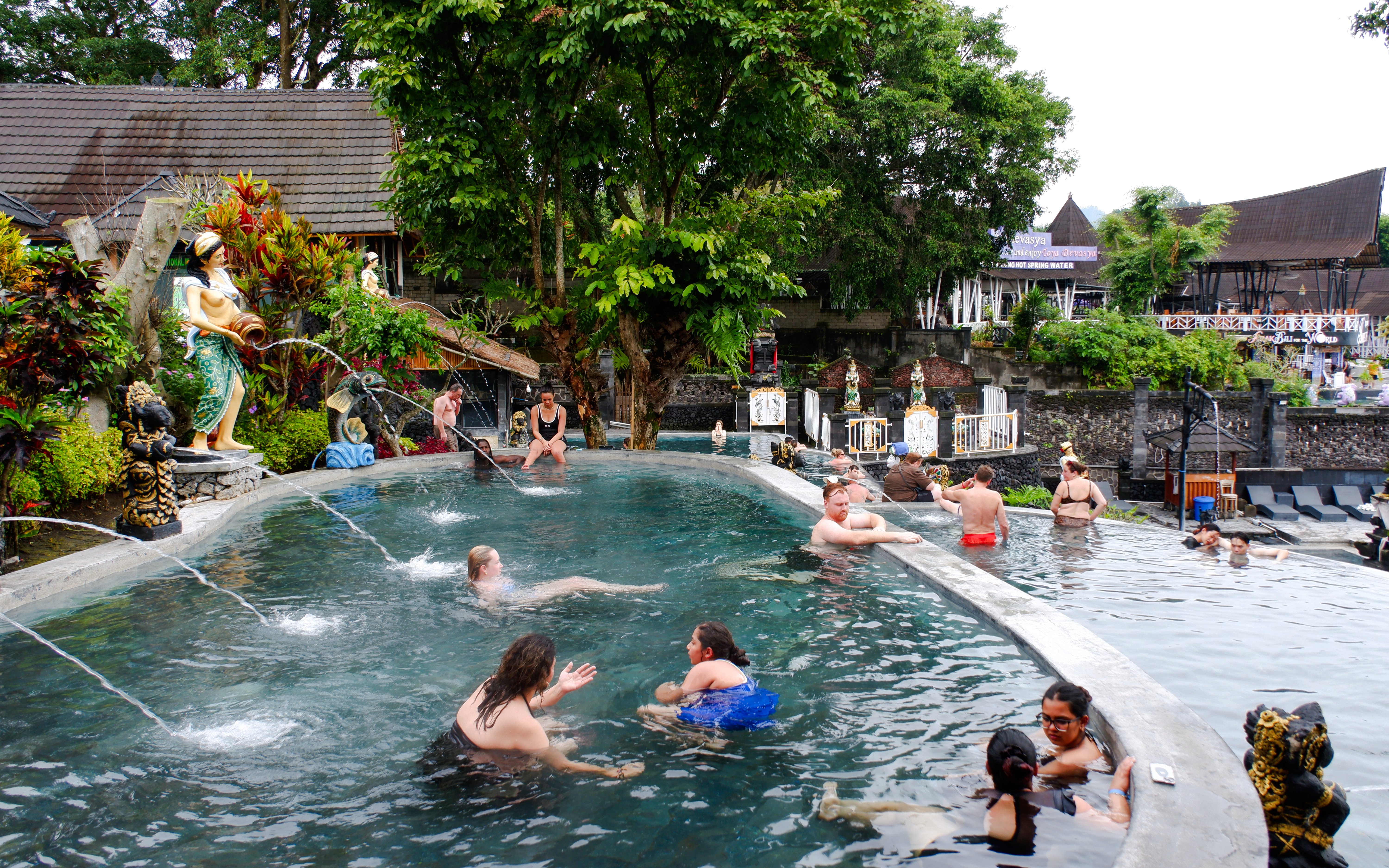Visitors enjoying a hot spring pool surrounded by statues and lush greenery in Indonesia.