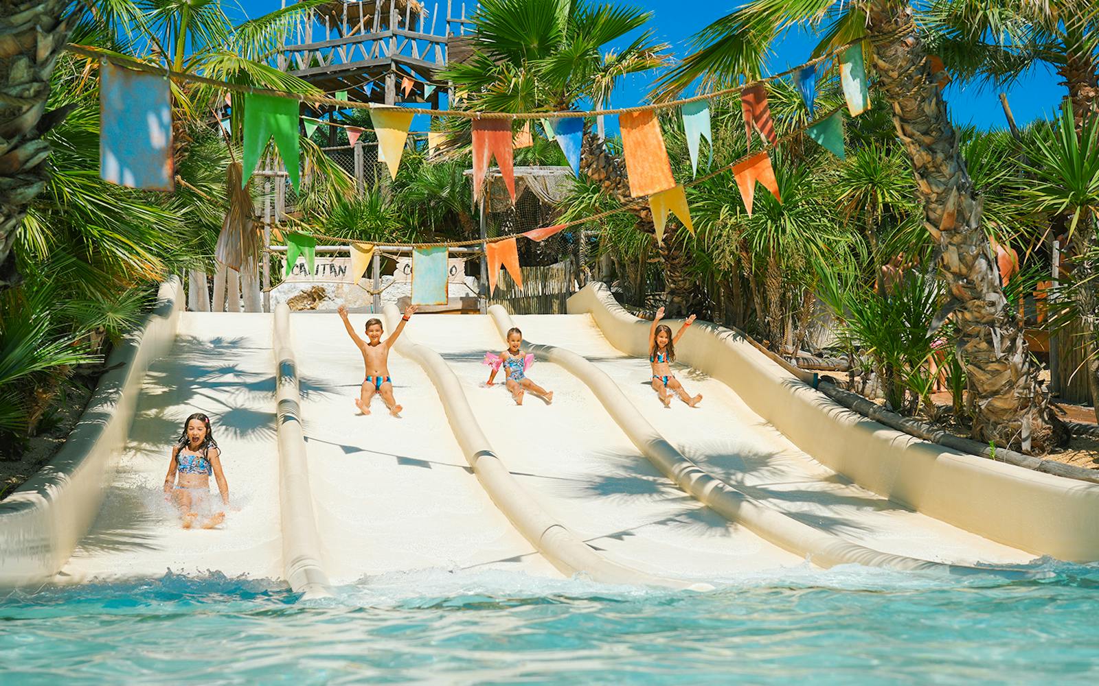 Kids sliding down water slides at Caribe Bay, surrounded by palm trees and colorful flags.