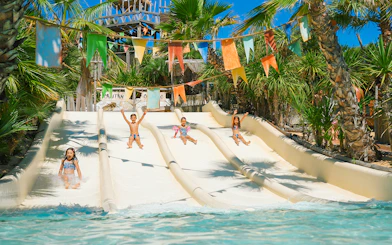 Kids sliding down water slides at Caribe Bay, surrounded by palm trees and colorful flags.