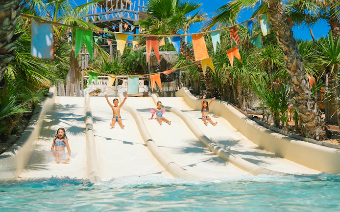 Kids sliding down water slides at Caribe Bay, surrounded by palm trees and colorful flags.