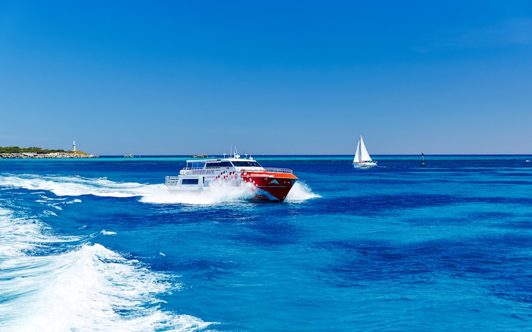 Ferry cruising on blue waters near Perth with a sailboat and lighthouse in the background.