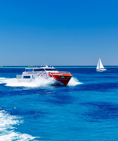 Ferry cruising on blue waters near Perth with a sailboat and lighthouse in the background.