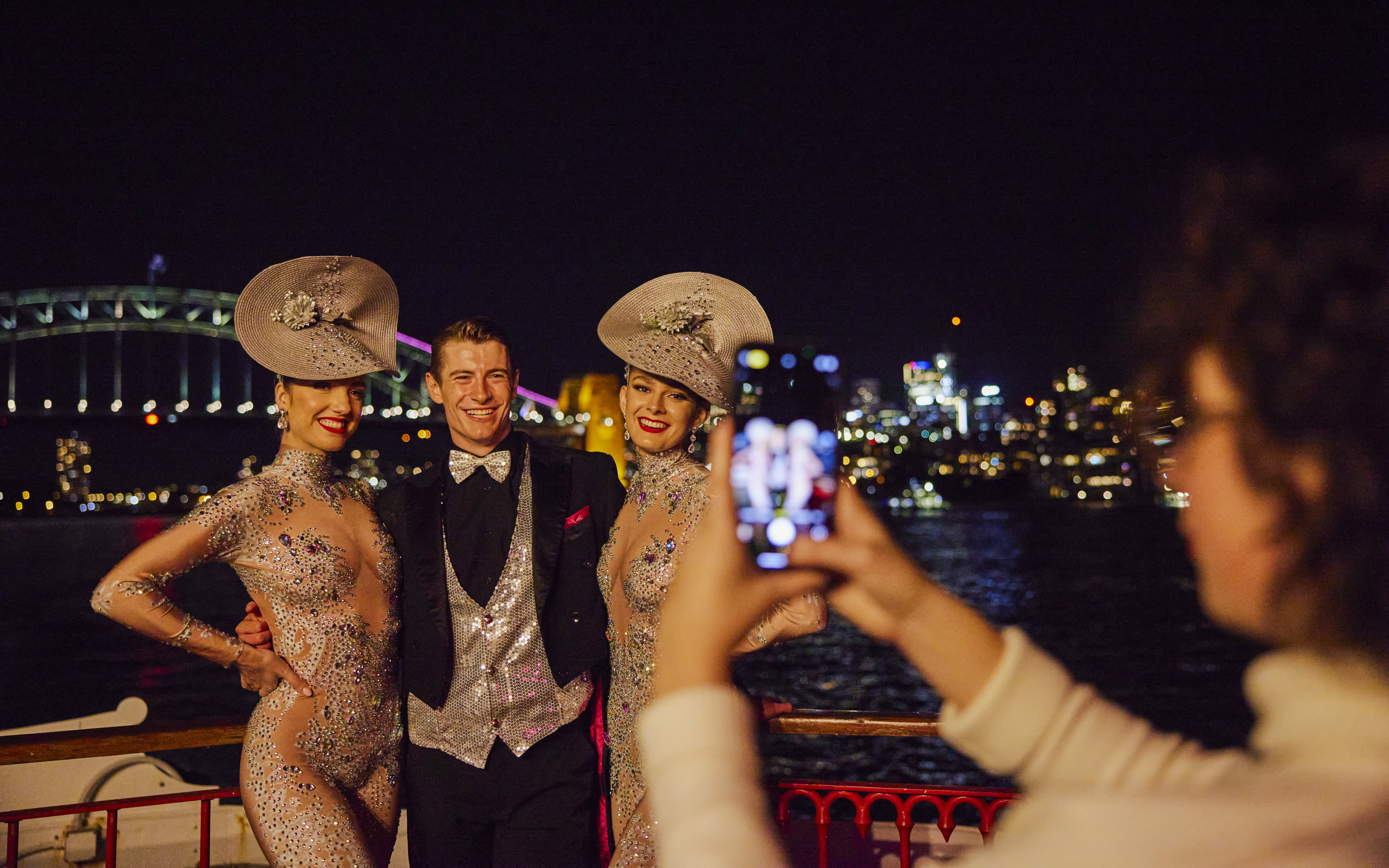 Cabaret performers pose on Showboat Vivid Sydney dinner cruise with Sydney Harbour Bridge in background.