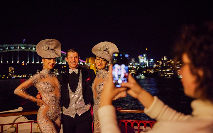 Cabaret performers pose on Showboat Vivid Sydney dinner cruise with Sydney Harbour Bridge in background.