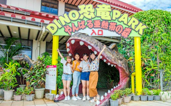 Visitors at Okashigoten Nago Dino Park entrance, standing by a dinosaur sculpture.