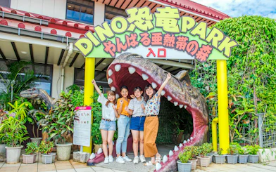 Visitors at Okashigoten Nago Dino Park entrance, standing by a dinosaur sculpture.