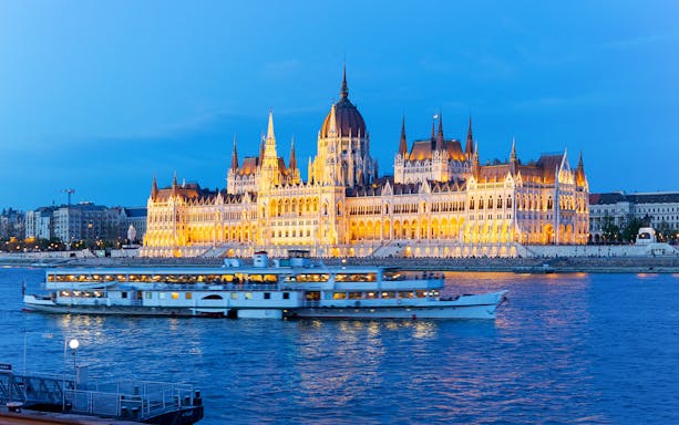 Cruise ship on Danube River with Budapest Parliament illuminated at dusk.