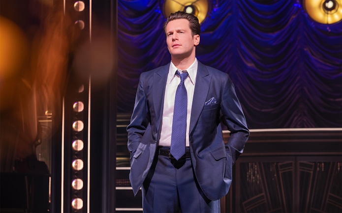 Man in a blue suit on stage with dramatic lighting and curtain backdrop.