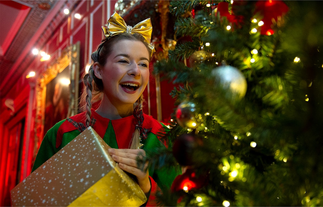 Person in festive attire holding a gift near a decorated Christmas tree at Warwick Castle.