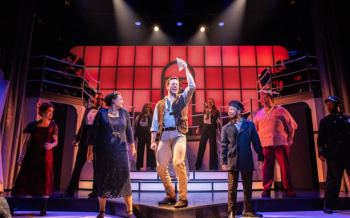 Cast performing on stage in the musical Titanique with vibrant red backdrop.