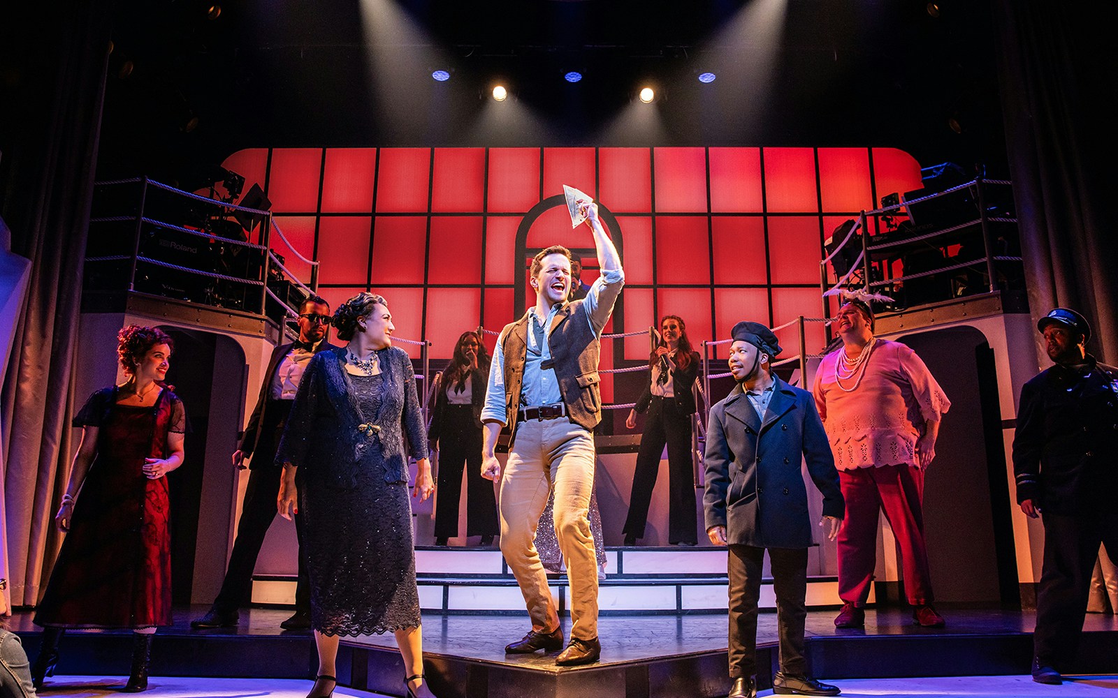 Cast performing on stage in the musical Titanique with vibrant red backdrop.
