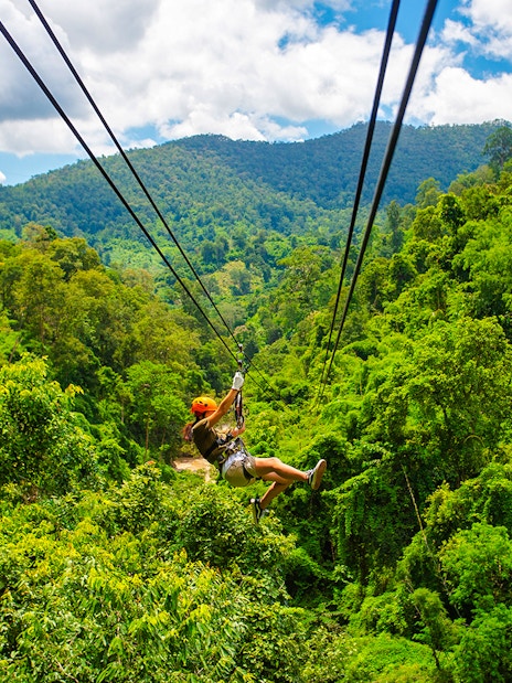 Person ziplining over lush green forest with mountains in the background.