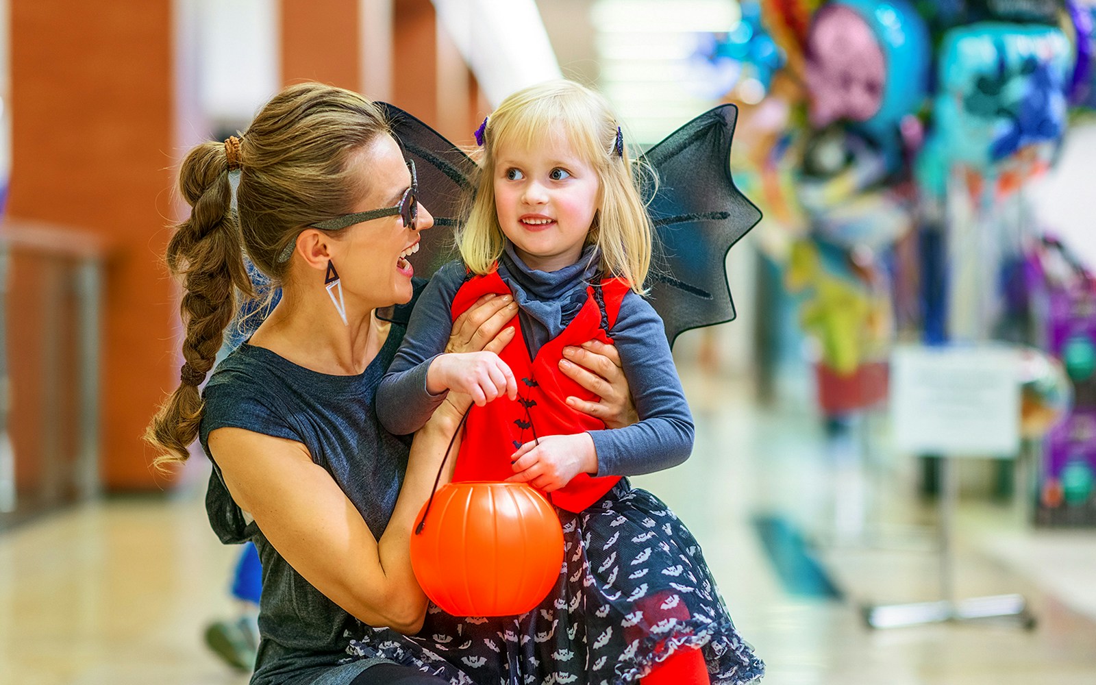 Mother and daughter in Halloween costumes at a mall, holding a pumpkin bucket.