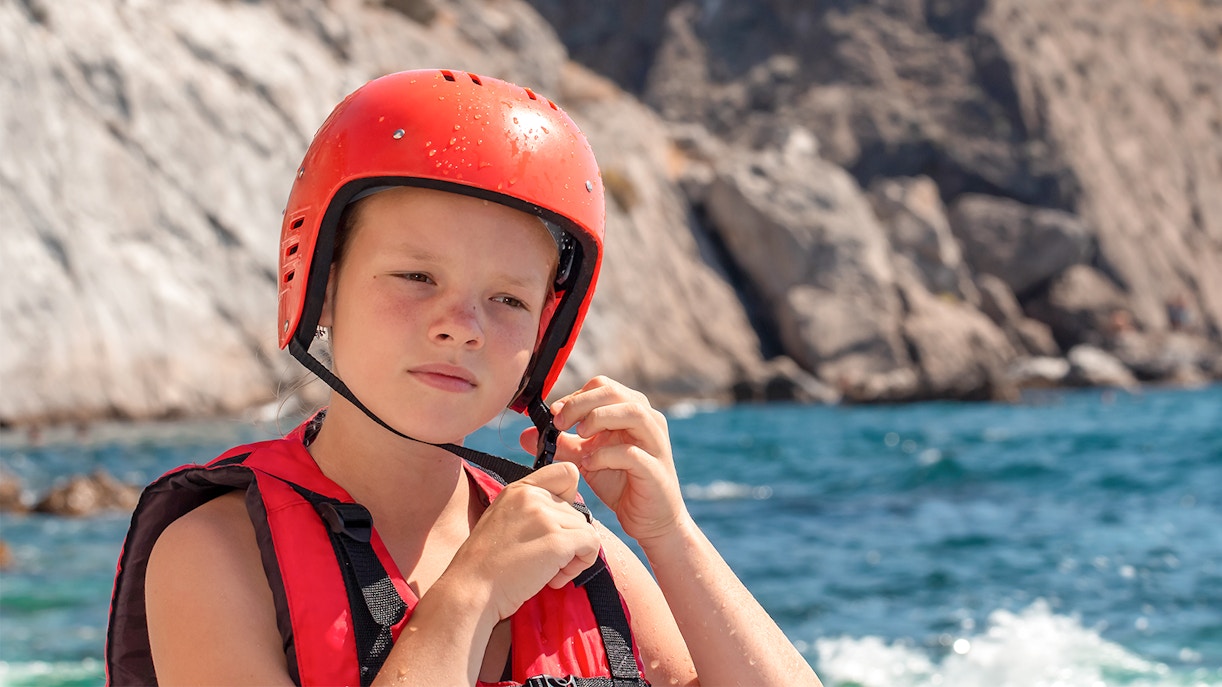 Girl in life jacket and red helmet on rocky seashore.