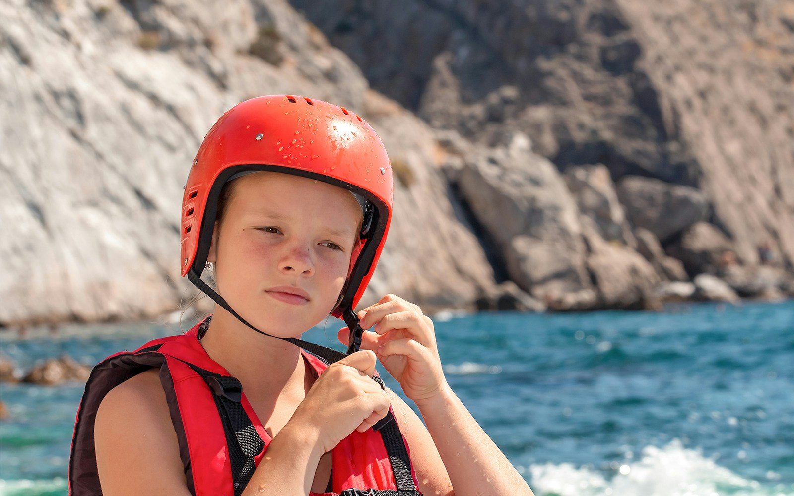 Girl in life jacket and red helmet on rocky seashore.