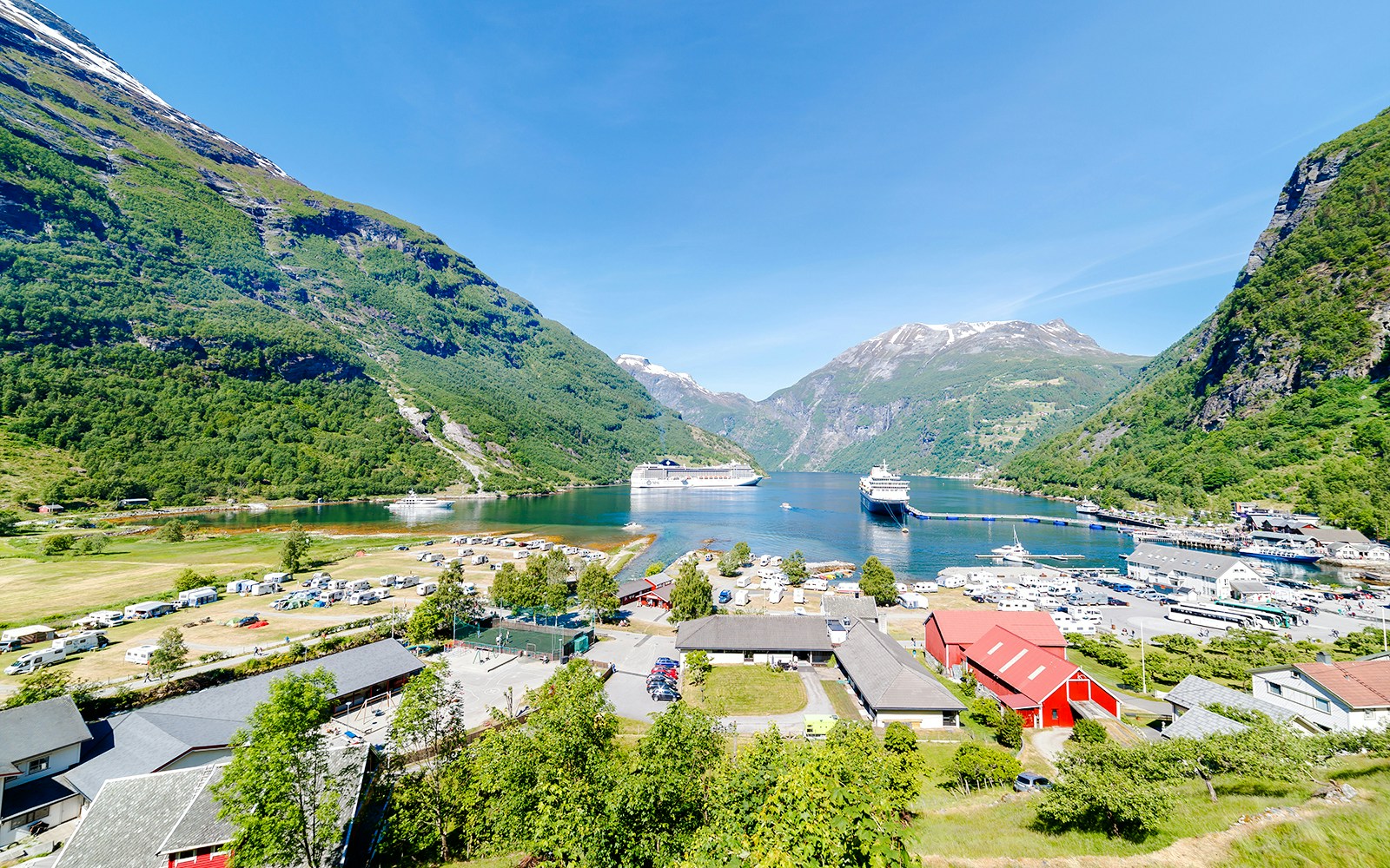Cruise ships docked at Norwegian fjord terminal surrounded by mountains and village.