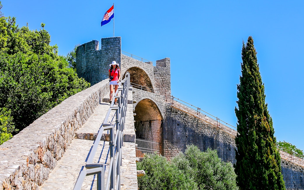 Young woman walking on the stone wall of Ston, Croatia, with a Croatian flag in the background.