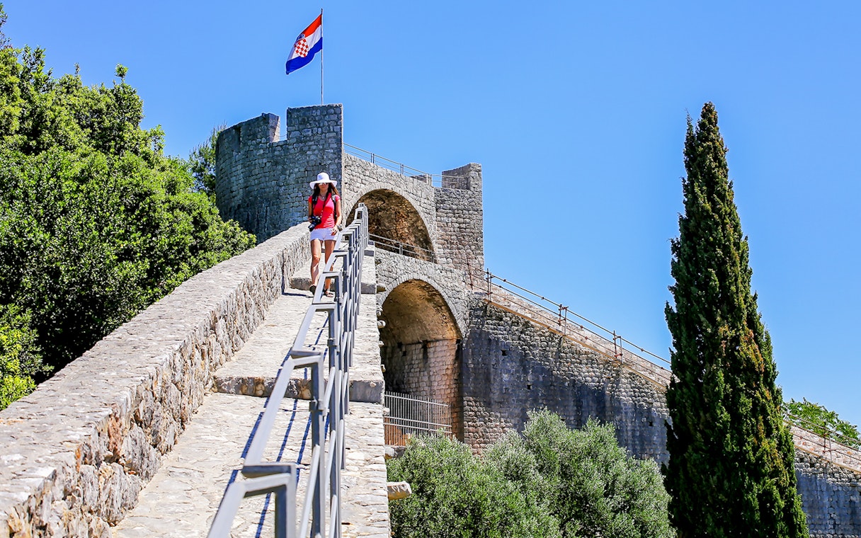 Young woman walking on the stone wall of Ston, Croatia, with a Croatian flag in the background.