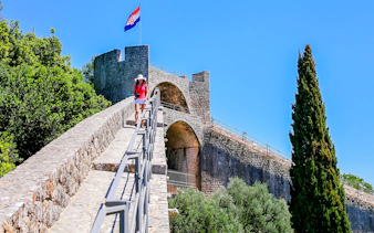 Young woman walking on the stone wall of Ston, Croatia, with a Croatian flag in the background.