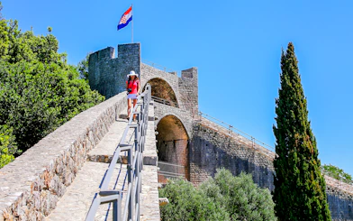 Young woman walking on the stone wall of Ston, Croatia, with a Croatian flag in the background.