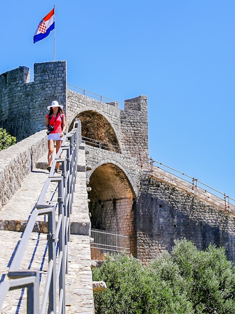 Young woman walking on the stone wall of Ston, Croatia, with a Croatian flag in the background.