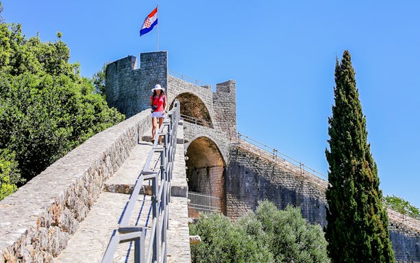 Young woman walking on the stone wall of Ston, Croatia, with a Croatian flag in the background.