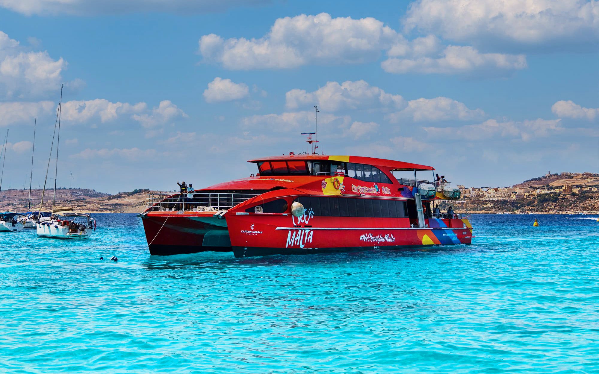 Red catamaran sailing in clear blue waters near Malta coast.