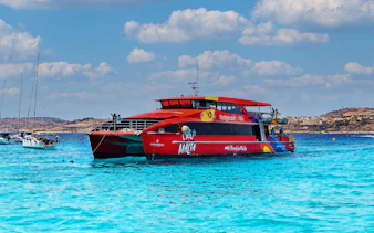Red catamaran sailing in clear blue waters near Malta coast.