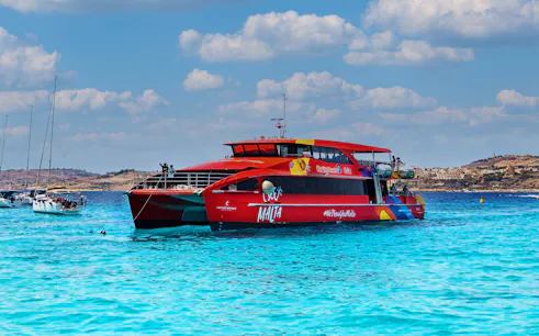 Red catamaran sailing in clear blue waters near Malta coast.
