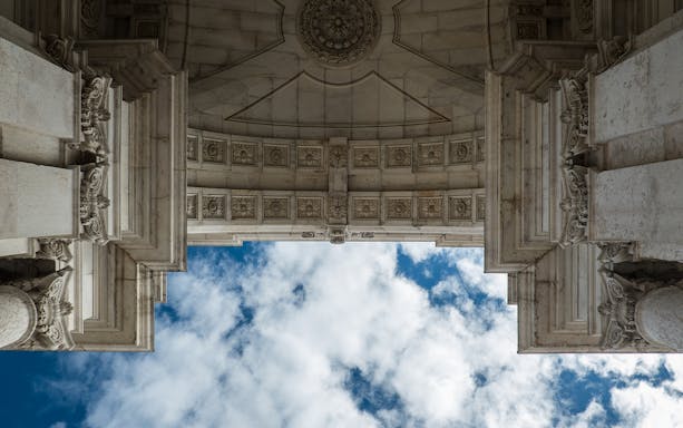 Looking up at the ornate archway of Rua Augusta Arch in Lisbon against a blue sky.