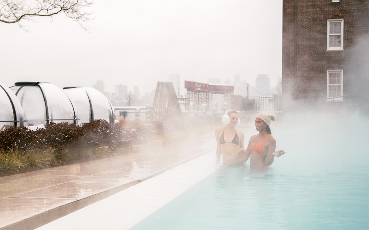 People enjoying a heated outdoor pool at QC New York with city skyline in the background.