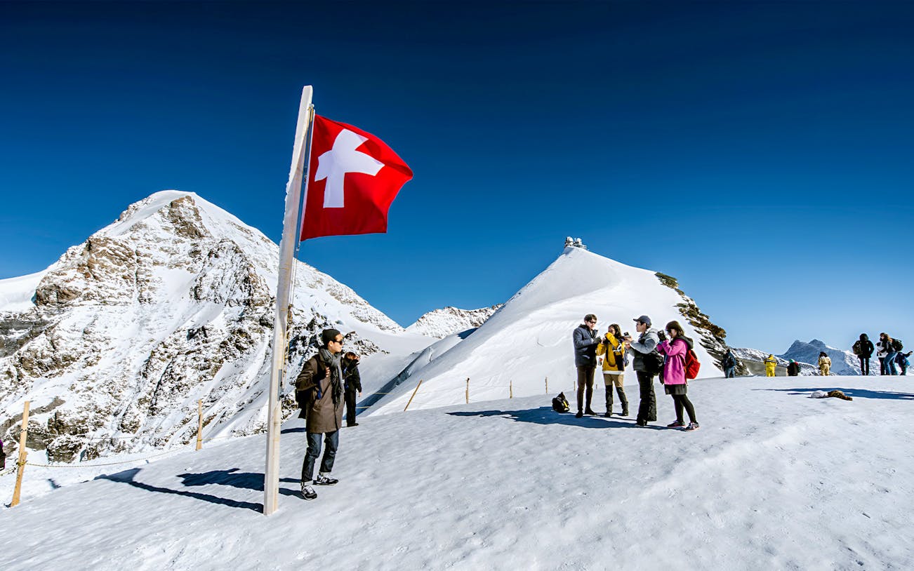 Tourists on snowy Schilthorn peak with Swiss flag, Interlaken day trip from Geneva.