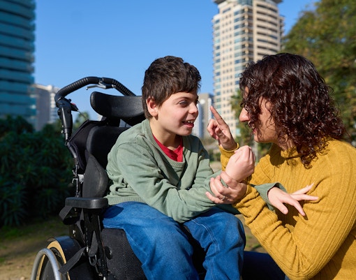 Disabled child in a wheelchair interacting with a tutor outdoors.