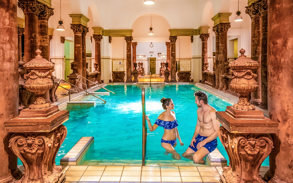 Indoor pool with ornate columns at Széchenyi Spa, Budapest, featuring two people enjoying the water.
