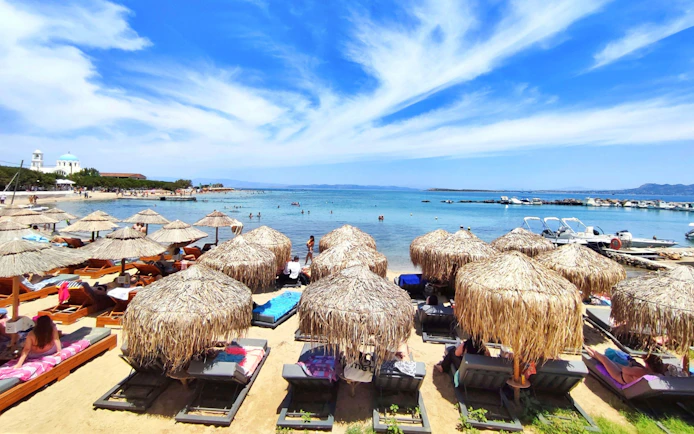 Sun loungers and straw umbrellas on Skala beach, Agistri island, Greece, with clear blue sea.