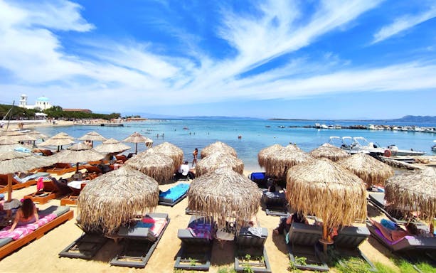 Sun loungers and straw umbrellas on Skala beach, Agistri island, Greece, with clear blue sea.