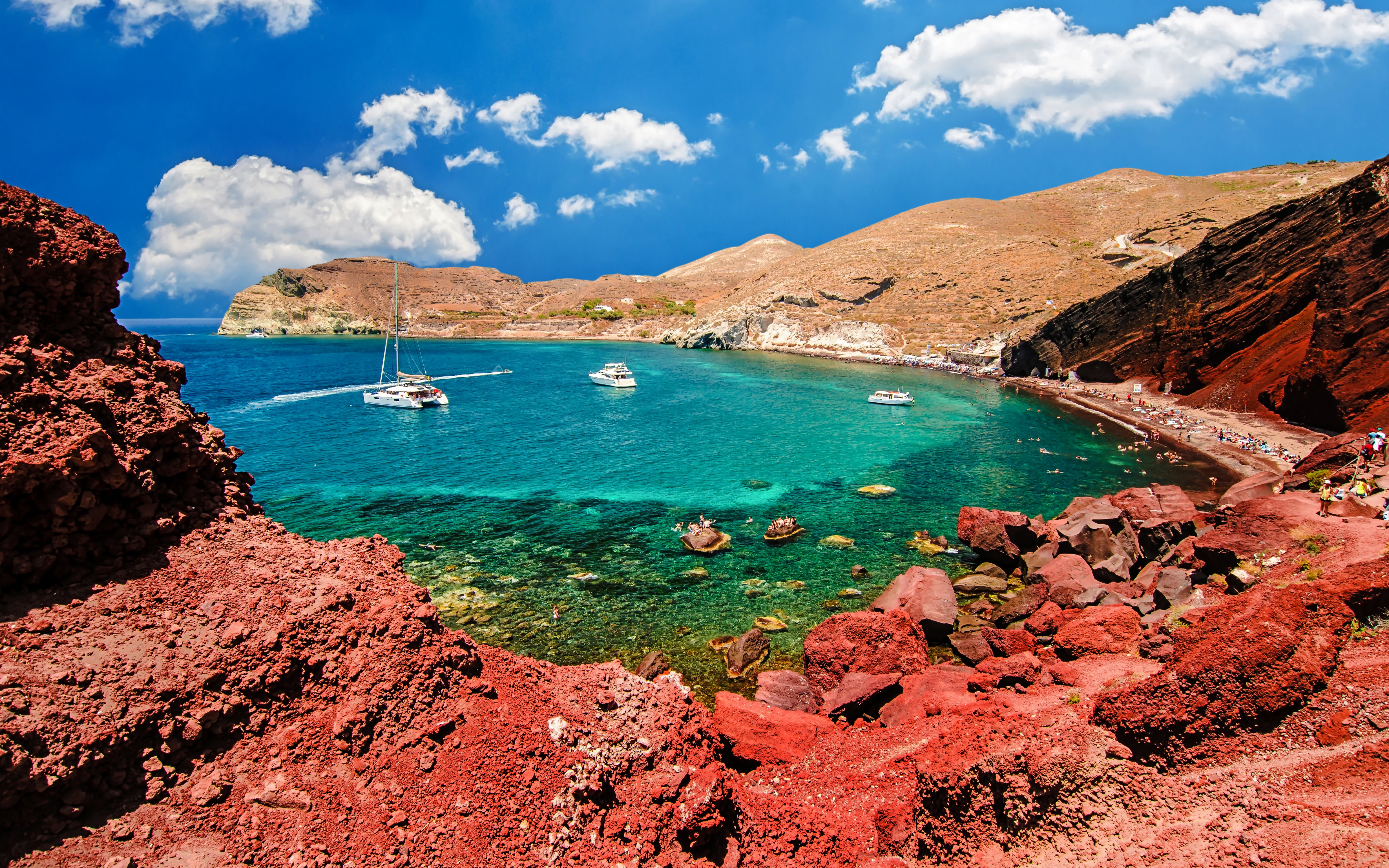 Catamarans on turquoise waters near Red Beach, Santorini, with red cliffs and tourists on the shore.