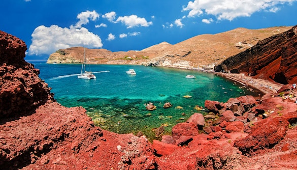 Catamarans on turquoise waters near Red Beach, Santorini, with red cliffs and tourists on the shore.